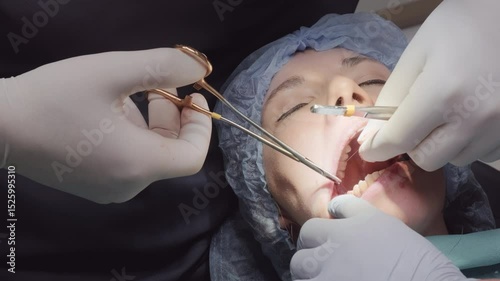 Doctor performs suturing in patient mouth using surgical forceps to manage thread and tissues. Assistant helps maintain access during wound closure