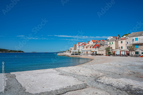 Fototapeta Naklejka Na Ścianę i Meble -  A stunning coastal town of Primosten in Croatia features traditional stone houses with red roofs beside the crystal-clear Adriatic Sea under a vibrant blue sky.