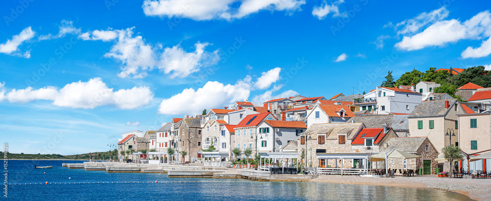 Fototapeta premium A stunning coastal town of Primosten in Croatia features traditional stone houses with red roofs beside the crystal-clear Adriatic Sea under a vibrant blue sky.