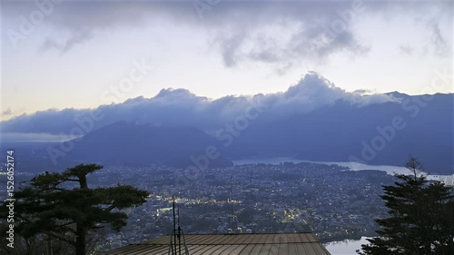 A breathtaking view of Fujikawaguchiko as twilight approaches, with distant mountains draped in clouds. The shimmering lake reflects the changing colors of the sky