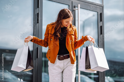 A woman with long hair smiles as she holds several shopping bags while standing outside a contemporary glass building on a sunny day. Consumer, fashion, shopping, shopping, lifestyle concept