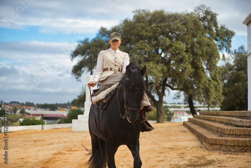 Young female horsewoman riding a black Spanish thoroughbred horse, trotting with him towards the camera. She has a saddle on the side and a riding crop. Concept horse riding, horsewoman, dressage.