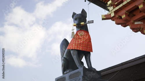 A black fox statue is dressed in a vibrant red garment, holding a decorative Japanese object in its mouth. Fushimi Inari, Kyoto, Japan