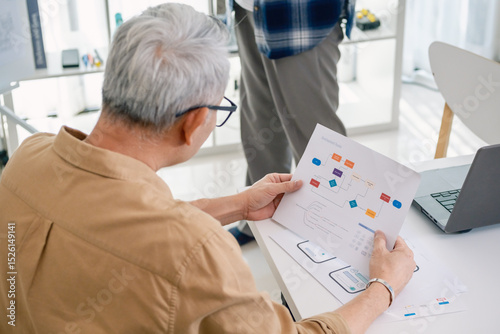 Senior programmer planning software development with flowchart and system process documentation at office