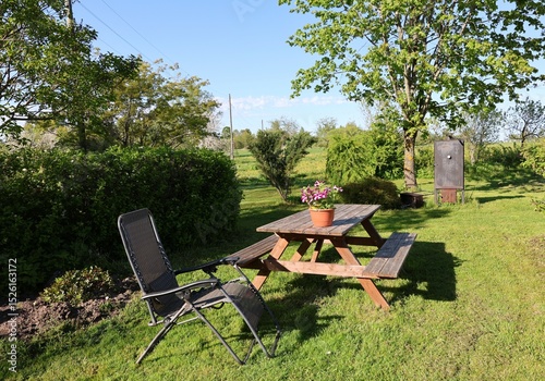 A peaceful garden scene with a wooden picnic table, lounge chair, and flower pot on a sunny lawn, surrounded by trees, hedges, and distant meadows under a clear blue sky.