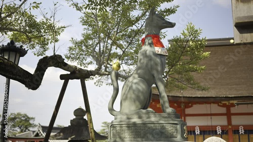 A graceful fox statue stands proudly at Fushimi Inari traditional Japanese shrine, decorated with a vibrant red scarf.