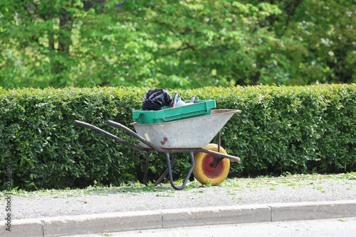 Rusty wheelbarrow with a yellow wheel and green crate full of garden tools rests beside a neatly trimmed hedge on a sunny day, symbolizing spring garden maintenance.