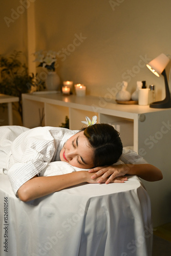 A woman is laying on a massage table with a white robe on. She is smiling and she is relaxed
