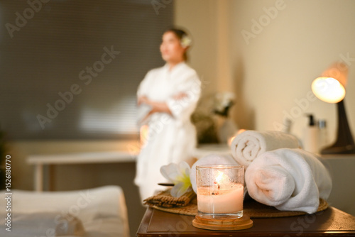 A woman is standing in front of a table with a candle and some towels. The candle is lit and the towels are neatly folded. The scene gives off a calm and relaxing atmosphere