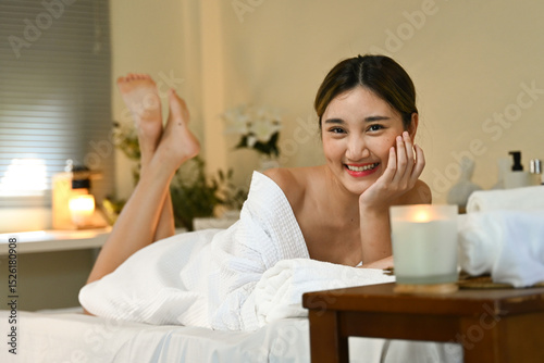 A woman is laying on a spa bed with a white robe on and a candle in front of her. She is smiling and she is enjoying her time.