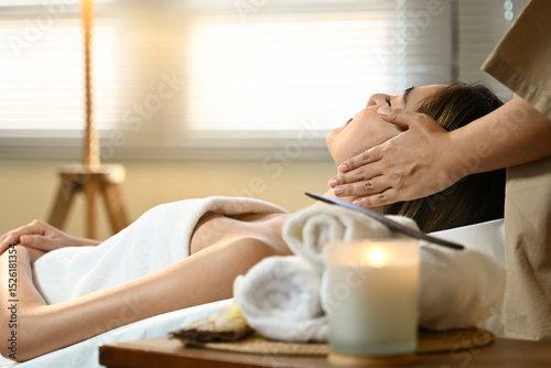 A woman is getting a massage while a massages therapist her head. The room is dimly lit and has candles and towels on the table