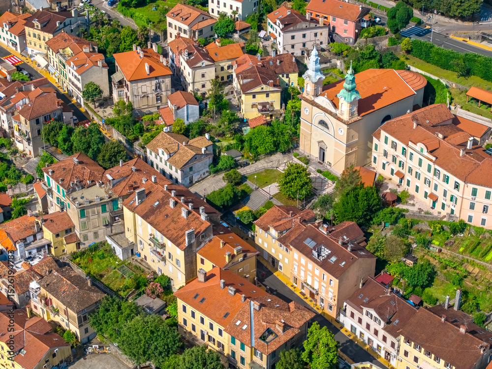 Obraz premium Aerial view of the central part of Volosko with a prominent twin-towered church, traditional Mediterranean houses, and green gardens, capturing the town’s authentic charm