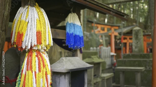 Brightly colored offerings hang at a shrine, showcasing threads of white, red, and blue. The tranquil atmosphere of the sacred space enhances the vivid decorations. Kyoto, Japan, Fushimi Inari