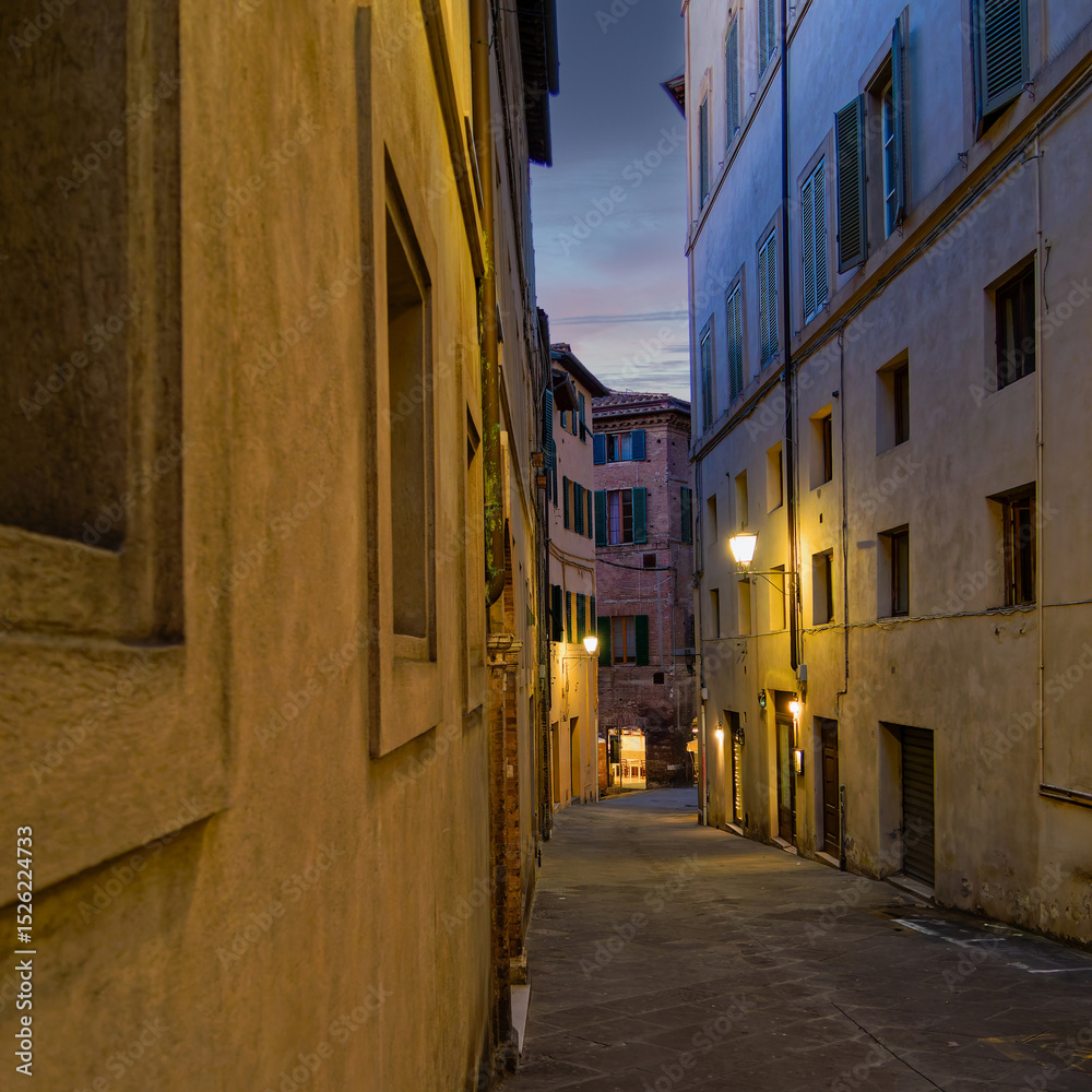 Fototapeta premium Twilight casts a warm glow over the narrow alleys of Siena, where historic stone buildings and elegant facades create an atmospheric and timeless urban scene. Travel to Tuscany, Italy.