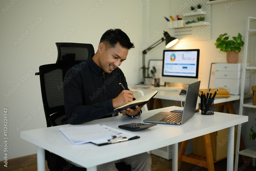 Fototapeta premium A Mature Asian businessman smiles while taking notes in a notebook at a modern office desk, with a laptop and documents around