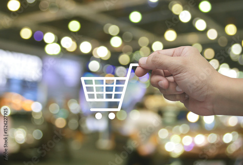 Hand holding shop cart flat icon in fingers over blur light and shadow of shopping mall, Business shopping online concept