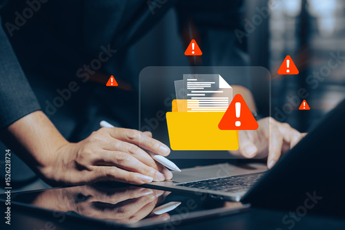 Digital data security breach alert: Close-up of hands using a laptop with a glowing orange warning icon on a file folder, surrounded by red exclamation marks.