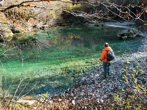 A man with a backpack, photographed from behind, stands by a forest lake with bright green water. A tourist during a visit to the Oko Skakavice (The Eye) location in Prokletije National Park