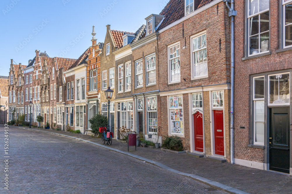 Obraz premium This captivating photo showcases a charming, historic street in Dordrecht, Netherlands, lined with classic Dutch gabled houses on a sunlit cobblestone path. Dordrecht, Netherlands. 16 March 2025.
