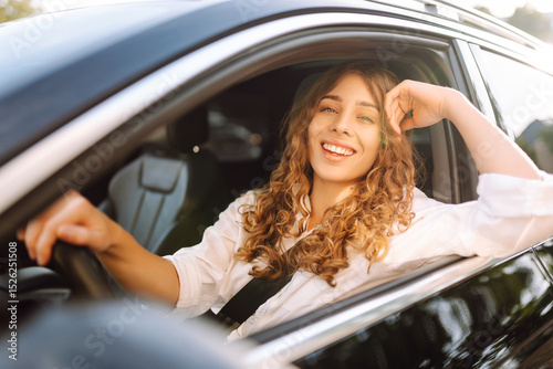 Beautiful woman driving a car in a white camisole is looking out the window. Young woman is driving a car and enjoying the sunny weather. Transportation, travel, taxi concept.