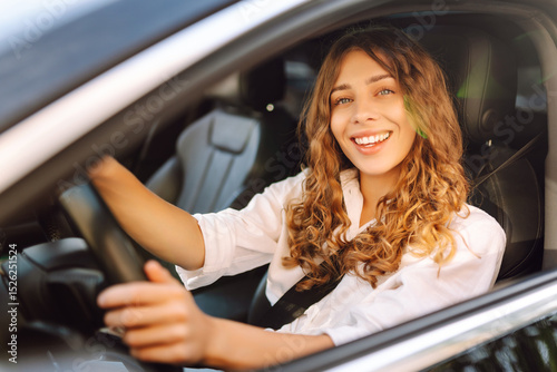 Beautiful woman driving a car in a white camisole is looking out the window. Young woman is driving a car and enjoying the sunny weather. Transportation, travel, taxi concept.