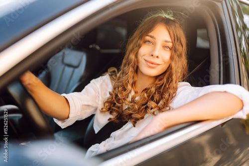 Beautiful woman driving a car in a white camisole is looking out the window. Young woman is driving a car and enjoying the sunny weather. Transportation, travel, taxi concept.