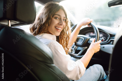 Beautiful woman driving a car in a white camisole is looking out the window. Young woman is driving a car and enjoying the sunny weather. Transportation, travel, taxi concept.