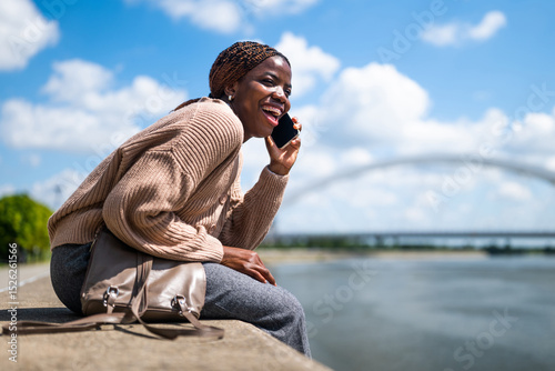 Young cheerful black woman is talking on the phone. She is laughing and enjoying sunny day in the city.