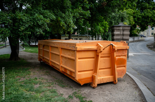 Schilderij op canvas Large orange industrial dumpster placed under trees in urban park area outdoors