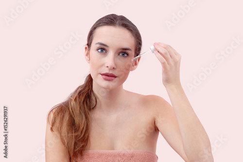 Woman applies serum to her face using a dropper. Radiating a sense of relaxation and self care. Isolated pink Background emphasizes the clean and fresh feeling associated with skincare routines