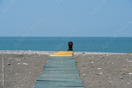 Fototapeta Naklejka Na Ścianę i Meble -  A single baby stroller on the beach of the Black Sea, Georgia. Wooden walkway to the beach with a stroller on it