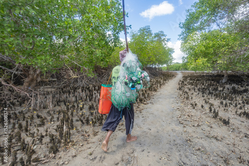 A fisherman walks barefoot through a lush mangrove forest carrying fishing nets and equipment, showcasing traditional methods of fishing amidst a natural coastal ecosystem under bright skies