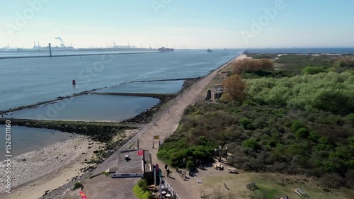 Aerial view of a coastal breakwater structure forming tidal pools, surrounded by greenish water and sandy shores. Waves gently touch the man-made barriers during low tide.