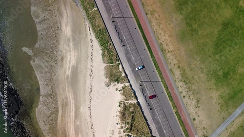 Aerial view of a coastal road with parked cars, a footpath through green grassland, and a small red lighthouse structure in a peaceful natural setting.