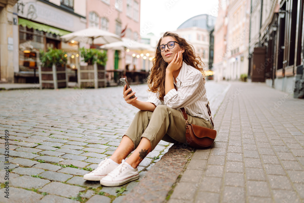 Fototapeta premium Portrait of a beautiful woman in glasses holding a phone on a sunset street. Beautiful woman texting on her phone, blogging and enjoying a walk in the city. Tourism concept.