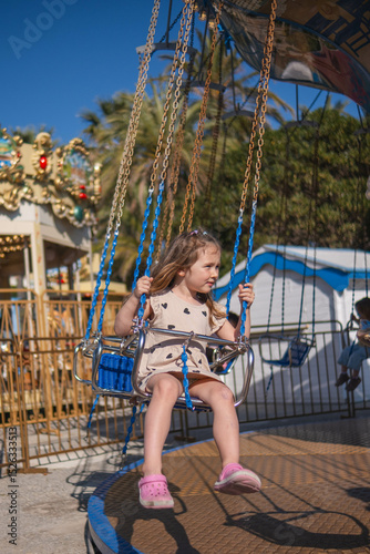 A little girl rides a carousel on a sunny summer day, laughing, waving and enjoying the rides. Palm trees grow in the background and other rides are visible