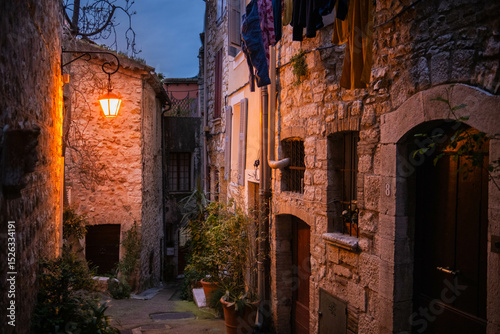 Fototapeta Naklejka Na Ścianę i Meble -  Evening old town Vence in France. Narrow cozy street with yellow vintage lantern that illuminates the road and clothes drying outside. Stone residential buildings