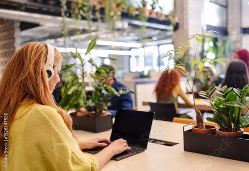 Young office worker wearing headphones typing on laptop in green workspace