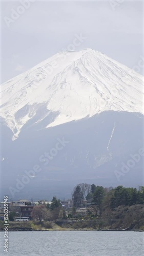 Mount Fuji rises magnificently, its snow-capped peak visible against a bright sky. The calm waters reflect the stunning mountain, surrounded by a picturesque village.