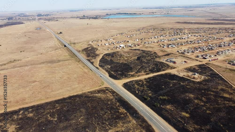Camera moving slowly of burned dry brown agricultural land and a settlement of small houses scattered next to a tarmac road 4K Aerial Video