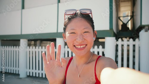 A cheerful young woman is happily waving at the camera while enjoying a bright sunny day at the beach, capturing a moment of joy and warmth in a picturesque coastal location.