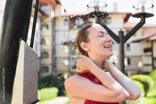 Happy woman in red swimsuit smiling under cold water in sunny hotel garden while taking outdoor shower