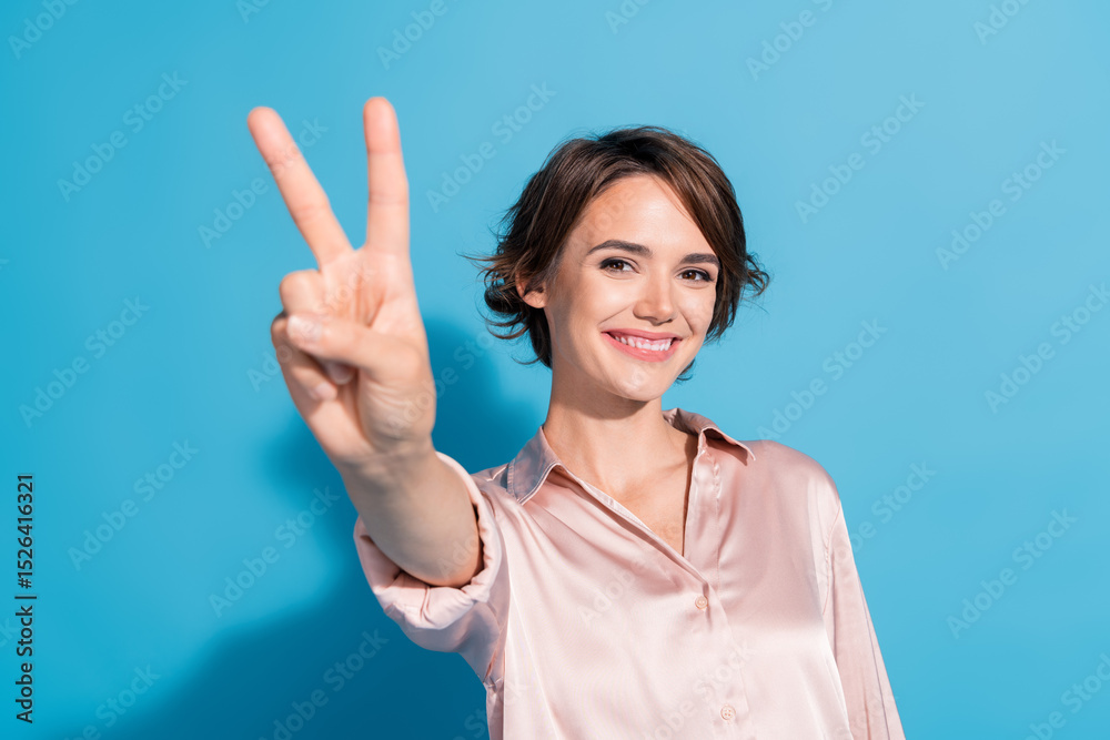 Confident young woman showing peace gesture with a bright smile against blue background, styled in satin pink blouse Confident young woman showing peace gesture with a bright smile against blue background, styled in satin pink blouse