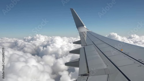 Aircraft wing soaring above pristine white cloud layer during bright sunny day, revealing extended ailerons and wing flaps against expansive blue sky backdrop