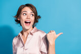 Young woman with stylish bob hairstyle in satin blouse gesturing with thumb and smiling joyfully against vibrant blue backdrop