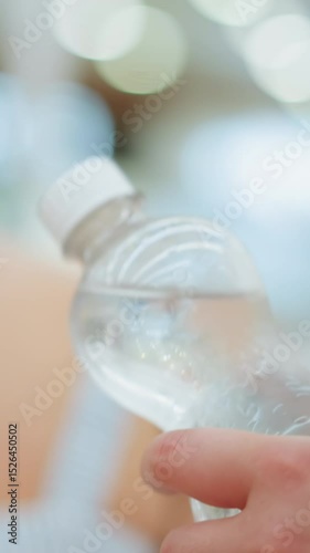 Lady holds chilled bottled water in a mall behind another woman, gradually shaking the bottle, the background features blurred lighting, and the bottle is clear and cold with visible condensation