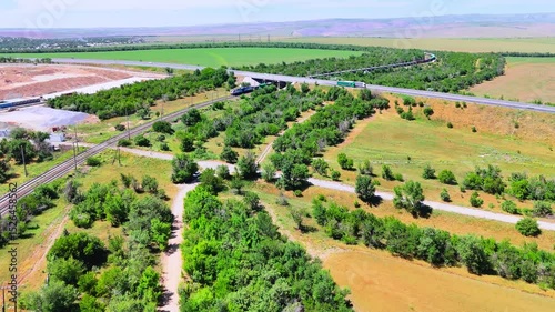 Green fields with a railway and highway stretching into the horizon. A freight train crosses the summer plains with mountains in the background.