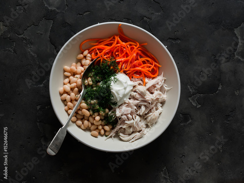 The idea of the salad - canned beans, boiled chicken fillet, spicy carrots, natural yogurt and dill in a bowl on a dark background, top view
