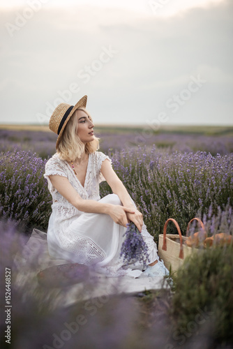 A serene scene of a woman in a white dress and straw hat seated amidst blooming lavender fields, holding a bundle of lavender flowers, enveloped by the calmness and beauty of nature. photo