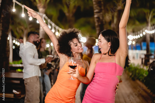 Two joyful women dancing with wine glasses during a fun tropical night party, surrounded by palm trees and string lights, celebrating friendship and enjoying the beach nightlife